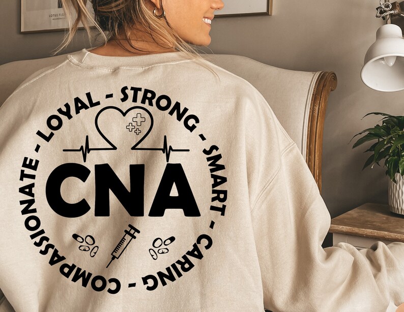 May include: A beige sweatshirt with a black circle design on the back. The design includes the words "CNA", "Loyal", "Strong", "Smart", "Caring", "Compassionate", and "Heart".