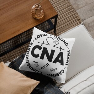 May include: White throw pillow with black text that reads "COMPASSIONATE - LOYAL - STRONG - SMART - CARING" in a circle around the word "CNA" with a heart beat line and a heart with a puzzle piece inside. There is a syringe and pills below the word "CNA".