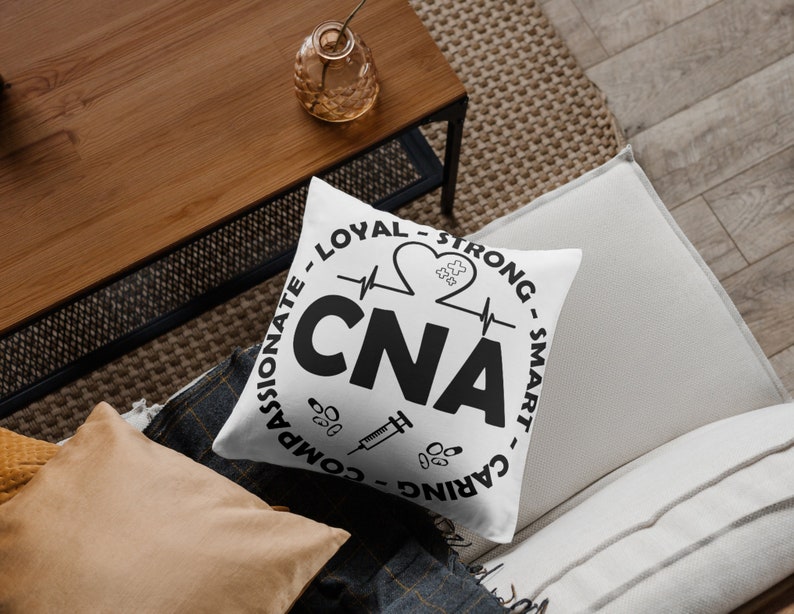 May include: White throw pillow with black text that reads "COMPASSIONATE - LOYAL - STRONG - SMART - CARING" in a circle around the word "CNA" with a heart beat line and a heart with a puzzle piece inside. There is a syringe and pills below the word "CNA".
