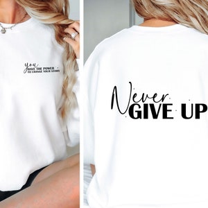 Op de afbeelding: Witte trui met de tekst "You have the power to change your story" op de voorkant en "Never Give Up!" op de achterkant.