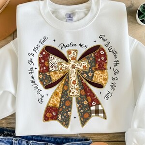 May include: White sweatshirt featuring a cross-shaped design with floral and plaid patterns. The design includes the text "God Is Within Her She Is Not Fall" and "Psalm 46:5". The sweatshirt has a crew neck and long sleeves.