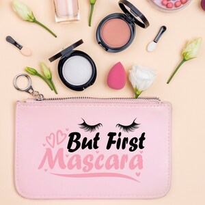 May include: Pink zippered pouch with the text "But First Mascara" in black and pink lettering. The pouch is surrounded by makeup products, including a blush compact, a foundation compact, and a makeup sponge.