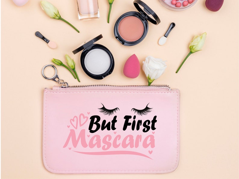 May include: Pink zippered pouch with the text "But First Mascara" in black and pink lettering. The pouch is surrounded by makeup products, including a blush compact, a foundation compact, and a makeup sponge.