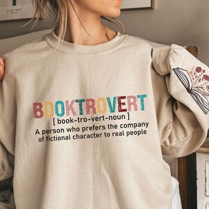 K&ouml;nnte beinhalten: Ein beiger Pullover mit dem Text "BOOKTROVERT [book-tro-vert-noun] A person who prefers the company of fictional character to real people" in bunten Buchstaben. Der Pullover hat ein Buch mit Blumen auf dem &Auml;rmel.