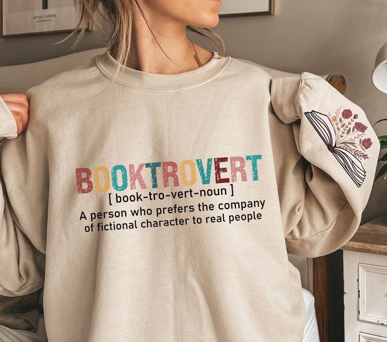K&ouml;nnte beinhalten: Ein beiger Pullover mit dem Text "BOOKTROVERT [book-tro-vert-noun] A person who prefers the company of fictional character to real people" in bunten Buchstaben. Der Pullover hat ein Buch mit Blumen auf dem &Auml;rmel.