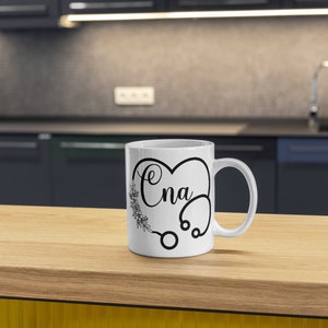May include: White ceramic coffee mug with a black design of a stethoscope in the shape of a heart with the word "CNA" inside.