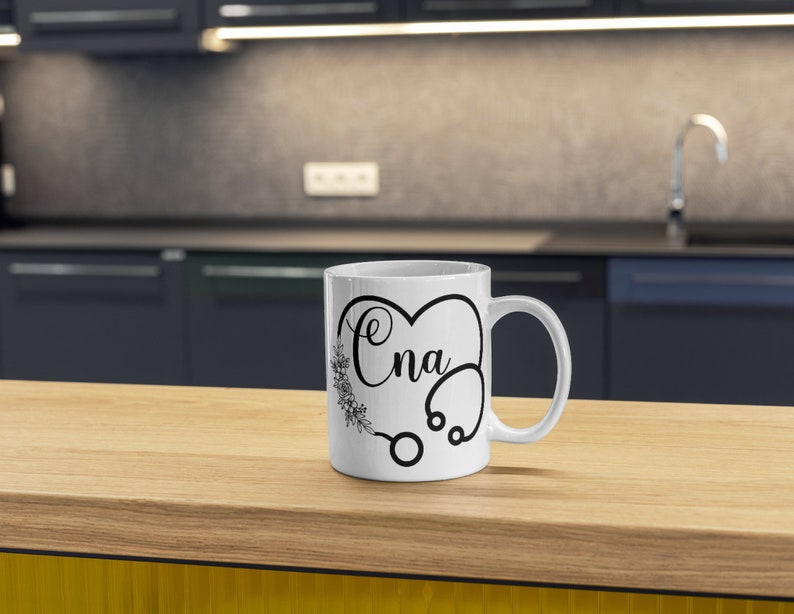 May include: White ceramic coffee mug with a black design of a stethoscope in the shape of a heart with the word "CNA" inside.