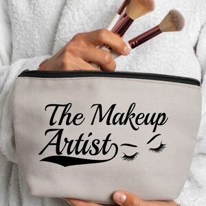 May include: A beige canvas makeup bag with a black zipper and the text "The Makeup Artist" in black cursive font with two stylized eyelashes on the right side.
