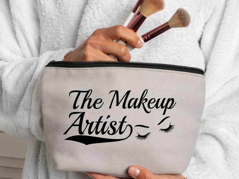 May include: A beige canvas makeup bag with a black zipper and the text "The Makeup Artist" in black cursive font with two stylized eyelashes on the right side.