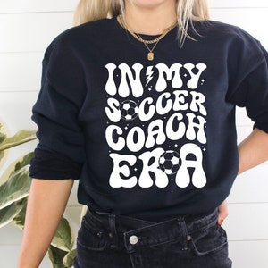 May include: Black sweatshirt with white text that reads "In My Soccer Coach Era" with a lightning bolt and soccer ball graphic.