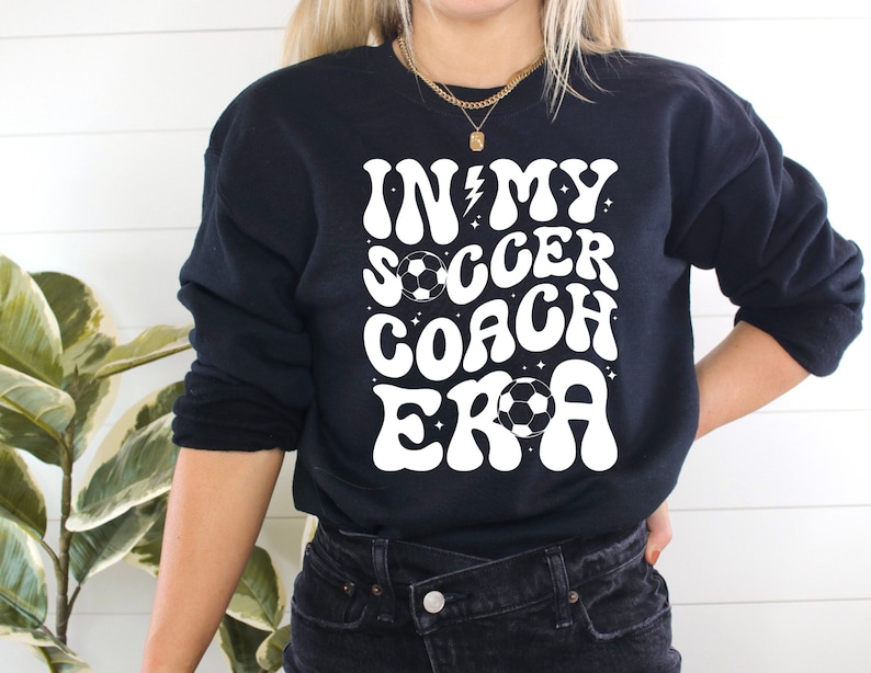 May include: Black sweatshirt with white text that reads "In My Soccer Coach Era" with a lightning bolt and soccer ball graphic.