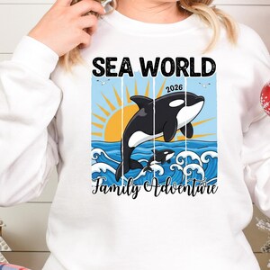 K&ouml;nnte beinhalten: Wei&szlig;es Sweatshirt mit einer Grafik, die einen schwarz-wei&szlig;en Orca-Wal zeigt, der &uuml;ber Ozeanwellen springt, mit einer untergehenden Sonne und den Worten "SEA WORLD 2026 Family Adventure".