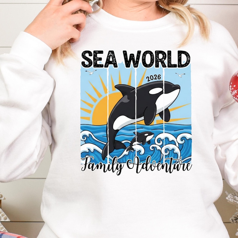 K&ouml;nnte beinhalten: Wei&szlig;es Sweatshirt mit einer Grafik, die einen schwarz-wei&szlig;en Orca-Wal zeigt, der &uuml;ber Ozeanwellen springt, mit einer untergehenden Sonne und den Worten "SEA WORLD 2026 Family Adventure".