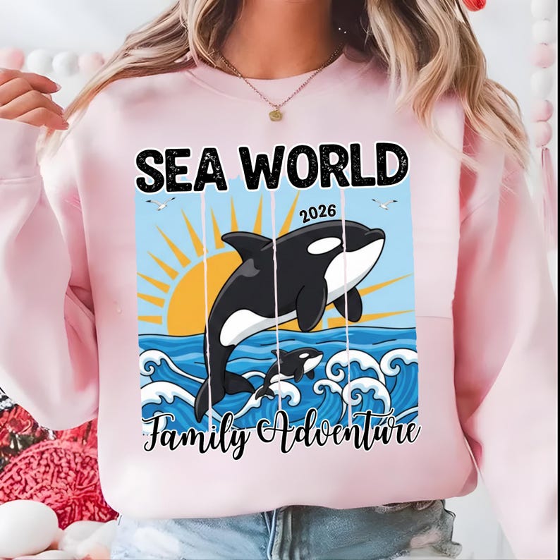 K&ouml;nnte beinhalten: Hellrosa Sweatshirt mit einer Grafik, die einen schwarz-wei&szlig;en Orca-Wal zeigt, der aus blauen Meereswellen springt. Die Worte "SEA WORLD 2026" und "Family Adventure" sind auf der Vorderseite aufgedruckt.