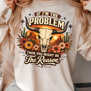 May include: Cream-colored sweatshirt featuring a graphic of a bull skull with long horns, set against a desert sunset scene with cacti and flowers. The text reads "If I'm the Problem Then You Might Be The Reason."