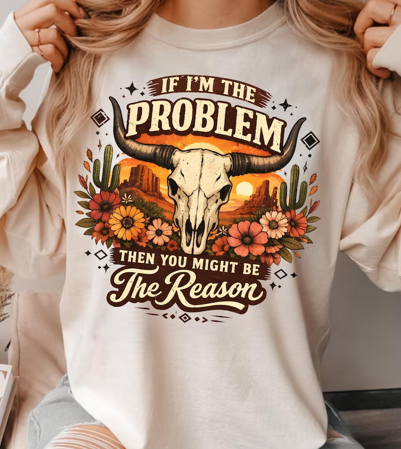 May include: Cream-colored sweatshirt featuring a graphic of a bull skull with long horns, set against a desert sunset scene with cacti and flowers. The text reads "If I'm the Problem Then You Might Be The Reason."