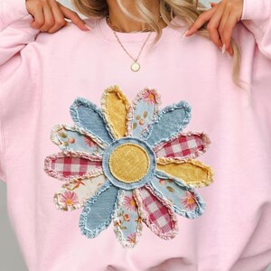 May include: A light pink sweatshirt featuring a large, appliqued daisy. The daisy has a yellow center and petals in various fabrics, including denim, floral prints, and gingham. The sweatshirt is a crew neck style.
