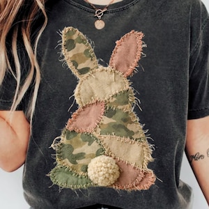 Può includere: T-shirt grigio scuro con un design a coniglio patchwork. Il coniglio è realizzato con vari tessuti, tra cui mimetico, beige e rosa, con una coda a pompon soffice. Il design è cucito sulla maglietta, creando un effetto strutturato.