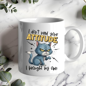 May include: White ceramic mug featuring a grumpy blue cat illustration with the text "I don't need your ATTITUDE, I brought my own." The cat has an angry expression and is surrounded by lightning bolt graphics.