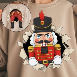 May include: Beige sweatshirt featuring a graphic of a cartoon nutcracker soldier bursting through a torn hole. The nutcracker is in a red uniform with a black hat and white gloves. A smaller image of the nutcracker's legs is on the back.