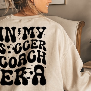May include: A beige sweatshirt with a black graphic that says "In My Soccer Coach Era" with a soccer ball graphic.