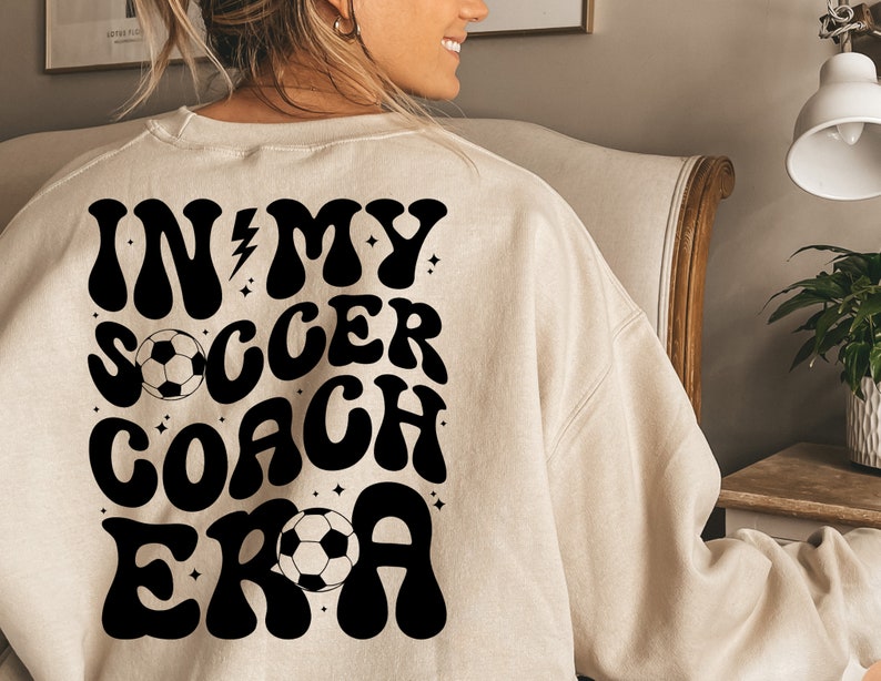 May include: A beige sweatshirt with a black graphic that says "In My Soccer Coach Era" with a soccer ball graphic.