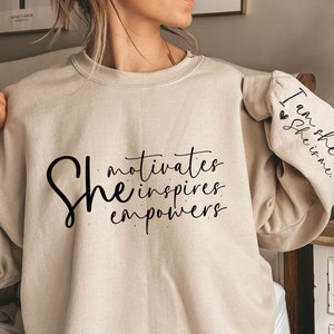 Może przedstawiać: Beżowa bluza z napisem "She motivates inspires empowers" z przodu i "I am she she is me" na rękawie.