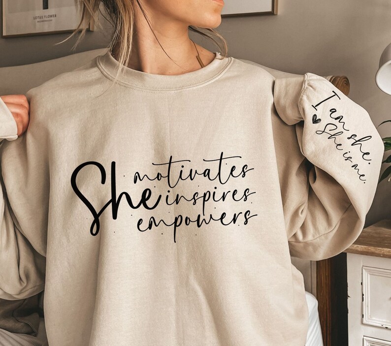 Może przedstawiać: Beżowa bluza z napisem "She motivates inspires empowers" z przodu i "I am she she is me" na rękawie.