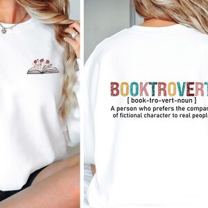 K&ouml;nnte beinhalten: Wei&szlig;er Pullover mit einem Buch- und Blumenmotiv auf der Vorderseite und dem Text "BOOKTROVERT [book-tro-vert-noun] A person who prefers the company of fictional character to real people" auf der R&uuml;ckseite.