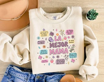 May include: A cream-colored sweatshirt with a graphic print of flowers and butterflies. The text on the sweatshirt reads "La Mejor Mama" in pink, purple, and blue lettering.