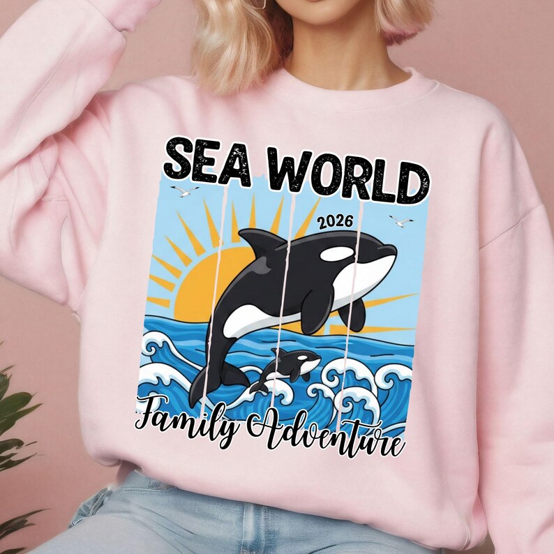 K&ouml;nnte beinhalten: Hellrosa Sweatshirt mit einer Grafik, die einen schwarz-wei&szlig;en Orca-Wal zeigt, der aus blauen Wellen unter einer gelben Sonne springt. Der Text "SEA WORLD 2026" und "Family Adventure" ist in schwarzer Schrift dargestellt.