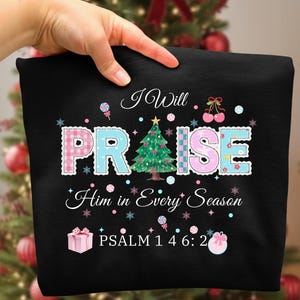 May include: Black sweatshirt with the words "I Will Praise Him in Every Season" in a decorative font. The word "PRAISE" is in pink and blue plaid letters with a Christmas tree in the center. Psalm 146:2 is printed at the bottom.