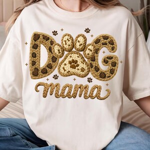 May include: Cream-colored t-shirt featuring the text "DOG mama" in a brown, textured design. The word "DOG" is stylized with paw prints and a leopard print pattern. Small paw prints and crosses are scattered around the text.
