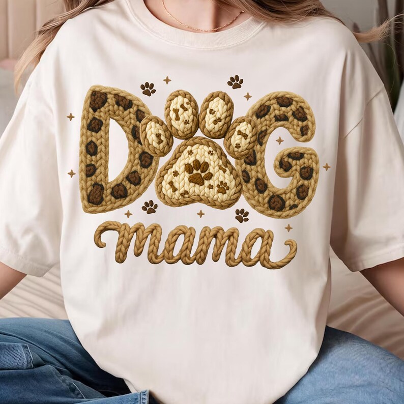 May include: Cream-colored t-shirt featuring the text "DOG mama" in a brown, textured design. The word "DOG" is stylized with paw prints and a leopard print pattern. Small paw prints and crosses are scattered around the text.