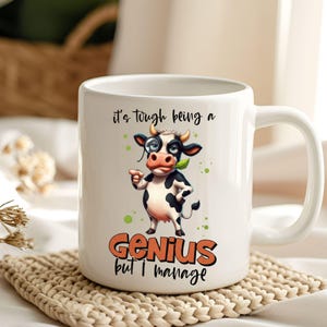 May include: White ceramic mug featuring a cartoon cow with a monocle and a leaf in its mouth. The mug has the text "it's tough being a Genius but I manage" in black and orange. The cow is black and white with green accents.
