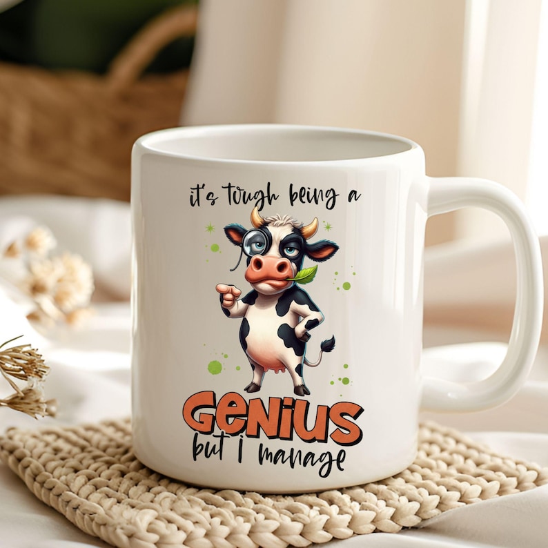 May include: White ceramic mug featuring a cartoon cow with a monocle and a leaf in its mouth. The mug has the text "it's tough being a Genius but I manage" in black and orange. The cow is black and white with green accents.