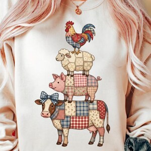 May include: Cream-coloured sweatshirt with a stacked farm animal design. A cow with a blue bow supports a pig, a sheep, and a rooster. Each animal is made of patchwork fabric in various colours.