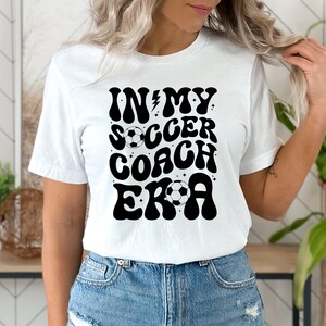 May include: White t-shirt with black text that reads "In My Soccer Coach Era" in a retro font. The text is decorated with stars and a soccer ball.