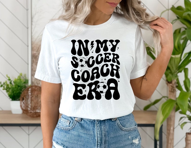 May include: White t-shirt with black text that reads "In My Soccer Coach Era" in a retro font. The text is decorated with stars and a soccer ball.