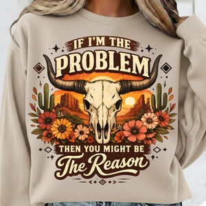 May include: Beige sweatshirt with a graphic featuring a bull skull, desert landscape, and floral accents. The text reads "IF I'M THE PROBLEM THEN YOU MIGHT BE The Reason". The design incorporates warm colors like orange, brown, and beige.