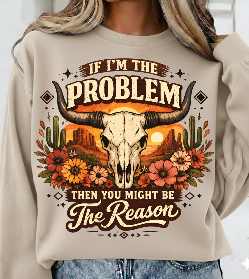 May include: Beige sweatshirt with a graphic featuring a bull skull, desert landscape, and floral accents. The text reads "IF I'M THE PROBLEM THEN YOU MIGHT BE The Reason". The design incorporates warm colors like orange, brown, and beige.