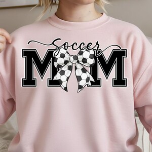May include: A light pink sweatshirt with the words "Soccer Mom" in black script and block letters. The word "MOM" is in large black block letters with a soccer ball bow in the center.