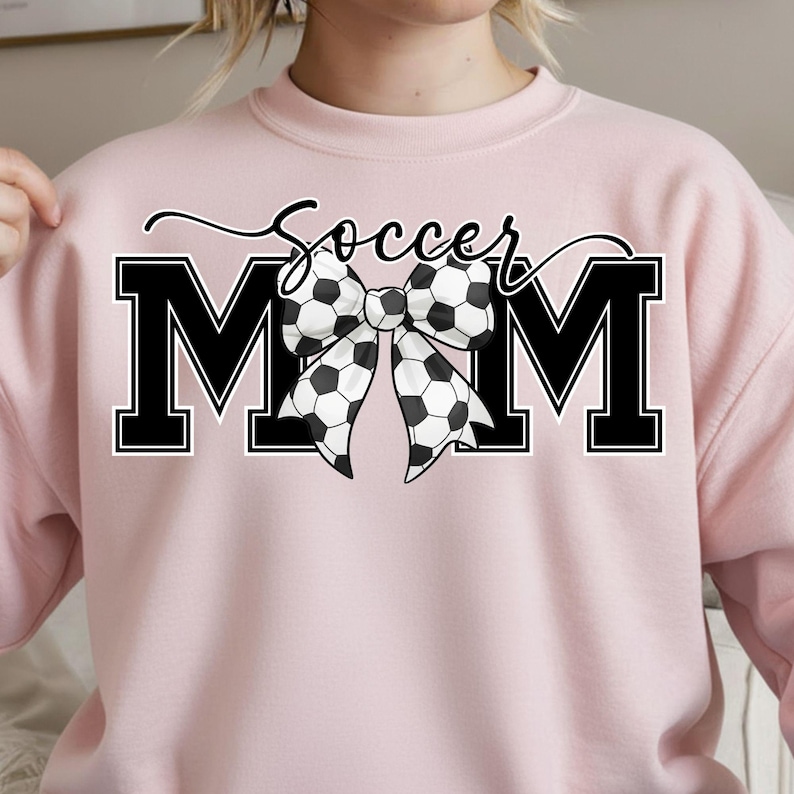 May include: A light pink sweatshirt with the words "Soccer Mom" in black script and block letters. The word "MOM" is in large black block letters with a soccer ball bow in the center.