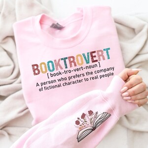 K&ouml;nnte beinhalten: Rosafarbener Pullover mit dem Text "BOOKTROVERT [book-tro-vert-noun] A person who prefers the company of fictional character to real people" und einem floralen Design eines offenen Buches auf dem &Auml;rmel.