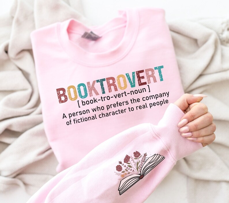 K&ouml;nnte beinhalten: Rosafarbener Pullover mit dem Text "BOOKTROVERT [book-tro-vert-noun] A person who prefers the company of fictional character to real people" und einem floralen Design eines offenen Buches auf dem &Auml;rmel.