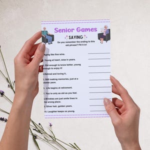 Peut inclure: Une feuille de jeu blanche intitul&eacute;e "Senior Games SAYING" avec des phrases &agrave; compl&eacute;ter. La feuille est tenue par deux mains et pr&eacute;sente des illustrations de personnes &acirc;g&eacute;es. Des brins de lavande sont en arri&egrave;re-plan.