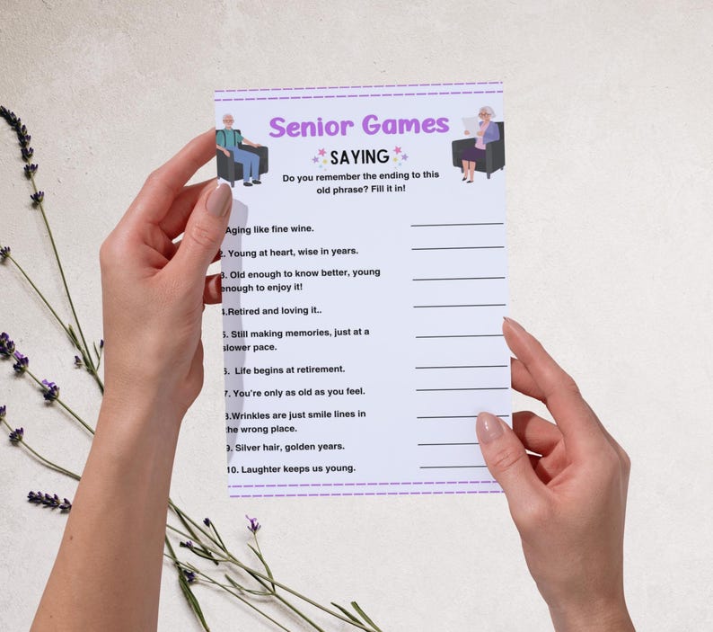 Peut inclure: Une feuille de jeu blanche intitul&eacute;e "Senior Games SAYING" avec des phrases &agrave; compl&eacute;ter. La feuille est tenue par deux mains et pr&eacute;sente des illustrations de personnes &acirc;g&eacute;es. Des brins de lavande sont en arri&egrave;re-plan.