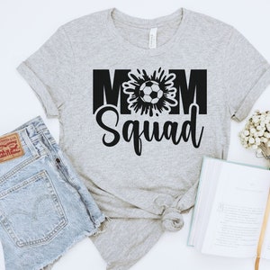 Puede incluir: Camiseta gris con texto negro que dice "Mom Squad" con un gr&aacute;fico de un bal&oacute;n de f&uacute;tbol en el medio de la palabra "Mom".