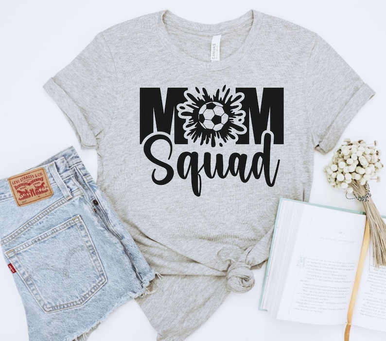 Puede incluir: Camiseta gris con texto negro que dice "Mom Squad" con un gr&aacute;fico de un bal&oacute;n de f&uacute;tbol en el medio de la palabra "Mom".