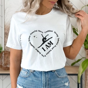 May include: White t-shirt featuring a black graphic of a gymnast within a heart shape. The words "I AM" are central, encircled by motivational terms such as "Driven," "Powerful," and "Confident."